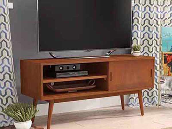 یک صفحه نمایشگر بزرگی روی یک میز زیر تلویزیونی چوبی به رنگ قهوه ای روشن قرار دارد و در کنار میز گلدان سفید رنگی می باشد و در داخل قفسه میز تلویزیون لوازم جانبی آن قرار گرفته است