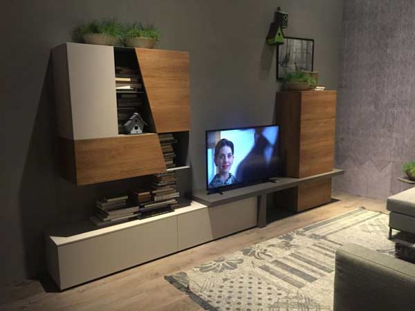 میز زیر تلویزیونی پایه ای همراه با قفسه هایی به صورت باز و بسته که متصل به دیوار می باشند در تصویر وجود دارد که رنگ دیوار پشتی آن طوسی و رنگ میز طوسی روشن همراه با تکه هایی از رنگ قهوه ای است