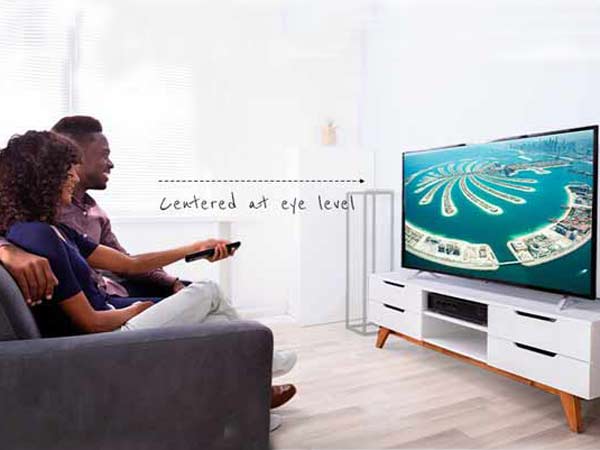 یک زن و مرد سیاه پوست بر روی کاناپه بنفش رنگی نشسته اند و به تی وی نگاه می کنند و صفحه نمایشگر به روی میز زیر تلویزیونی سفیدی قرار گرفته است.