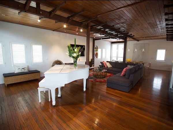 اتاق نشیمنی با کف و سقف چوبی به رنگ قهوه ای تیره و روشن است و پیانو سفیدی در آن قرار دارد و یک گلدان روی آن می باشد و مبل های راحتی قهوه ای تیره ای همراه با یک میز جلو مبلی در تصویر می باشد