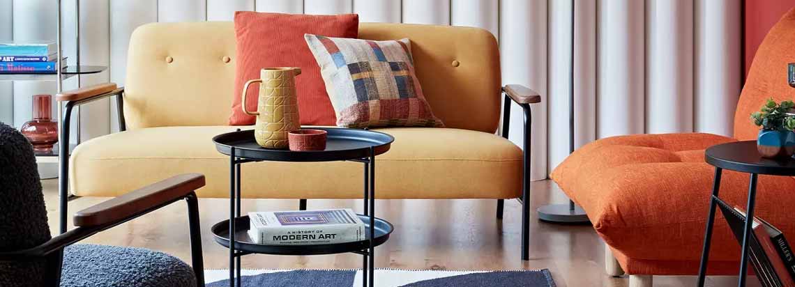 اتاقی با یک دست مبل با ترکیب رنگ نارنجی و زرد به همراه فرش سورمه ای سفید و صندلی سورمه ای و کتابخانه ای در گوشه آن