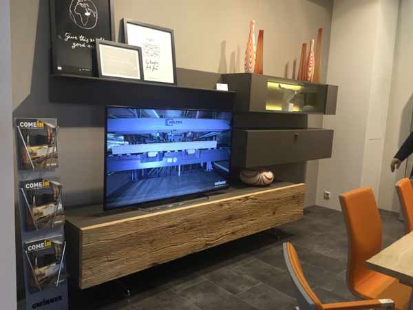 باکس تلویزیون چوبی قفسه باز با ترکیب رنگ های مشکی و قهوه ای روشن در تصویر می باشد که نمایشگر در مرکز آن قرار گرفته است و در بالای آن قفسه بازی می باشد که روی آن با استفاده از سه قاب تزیین شده است و رو به روی میز نمایشگر صندلی نارنجی رنگی وجود دارد.