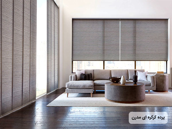 عکس پرده کرکره اي خاکستري رنگ روي يک ديوار بلند با پنجره بزرگ به همراه يک مبل ال شکل خاکستري و يک ميز دايره اي شکل از جنس چوب و پارچه .