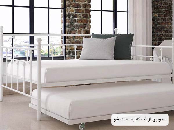 تصويری از يک مبل تختخوابشو مدرن به رنگ سفيد که دو عدد کوسن به رنگ های خاکستری و طوسی روی آن قرار گرفته اند.