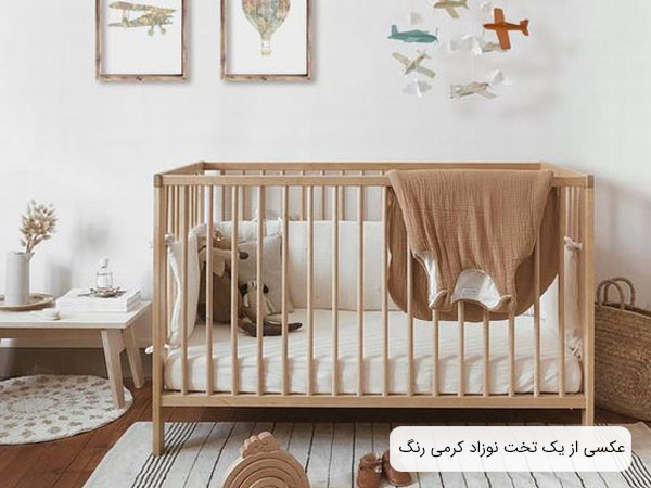عکسی از يک تخت خواب نوزاد کرمی رنگ در کنار اشيا و لوازمی با همين طيف رنگ و در يک اتاق با ديوار سفيد .