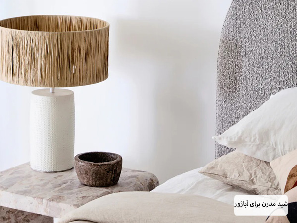 یک آباژور مدرن با بدنه سفید رنگ و شید دایره ای مدل چوبی در کنار تخت خواب.