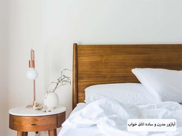 یک عدد اباژور رومیزی با طراحی مدرن و مینیمال بدون شید و با پایه های چوبی که کنار یک تخت خواب قرار گرفته است.